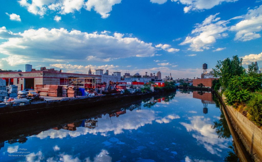 Clouds reflect in the water of the Gowanus Canal looking north with the Twin Towers peeking above the shoreline in 1989. The wall reflected in the water has no graffiti in 1989 and has now joined the past. ©Mark D Phillips